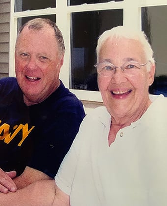 Stanley Rock (left) is smiling and wearing a dark shirt with yellow letters reading NAVY. Nancy Rock (right) is wearing a white blouse, glasses and smiling. 